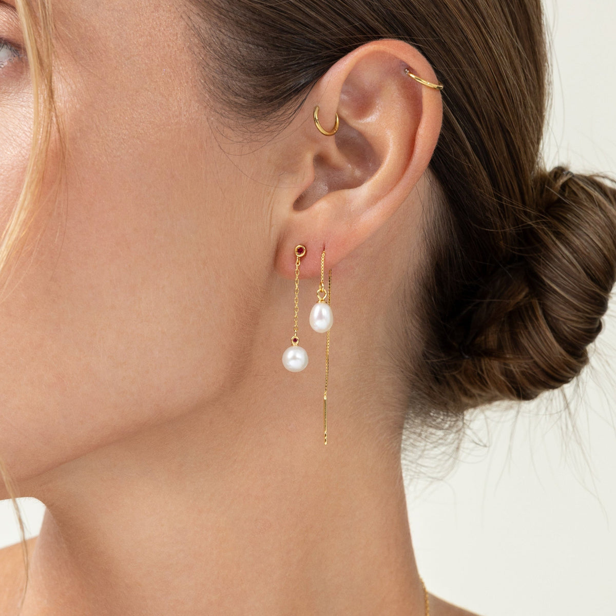 Model wearing an ear stack of freshwater pearl drop earrings, a small red lab-grown ruby stud, and gold hoops in the upper ear