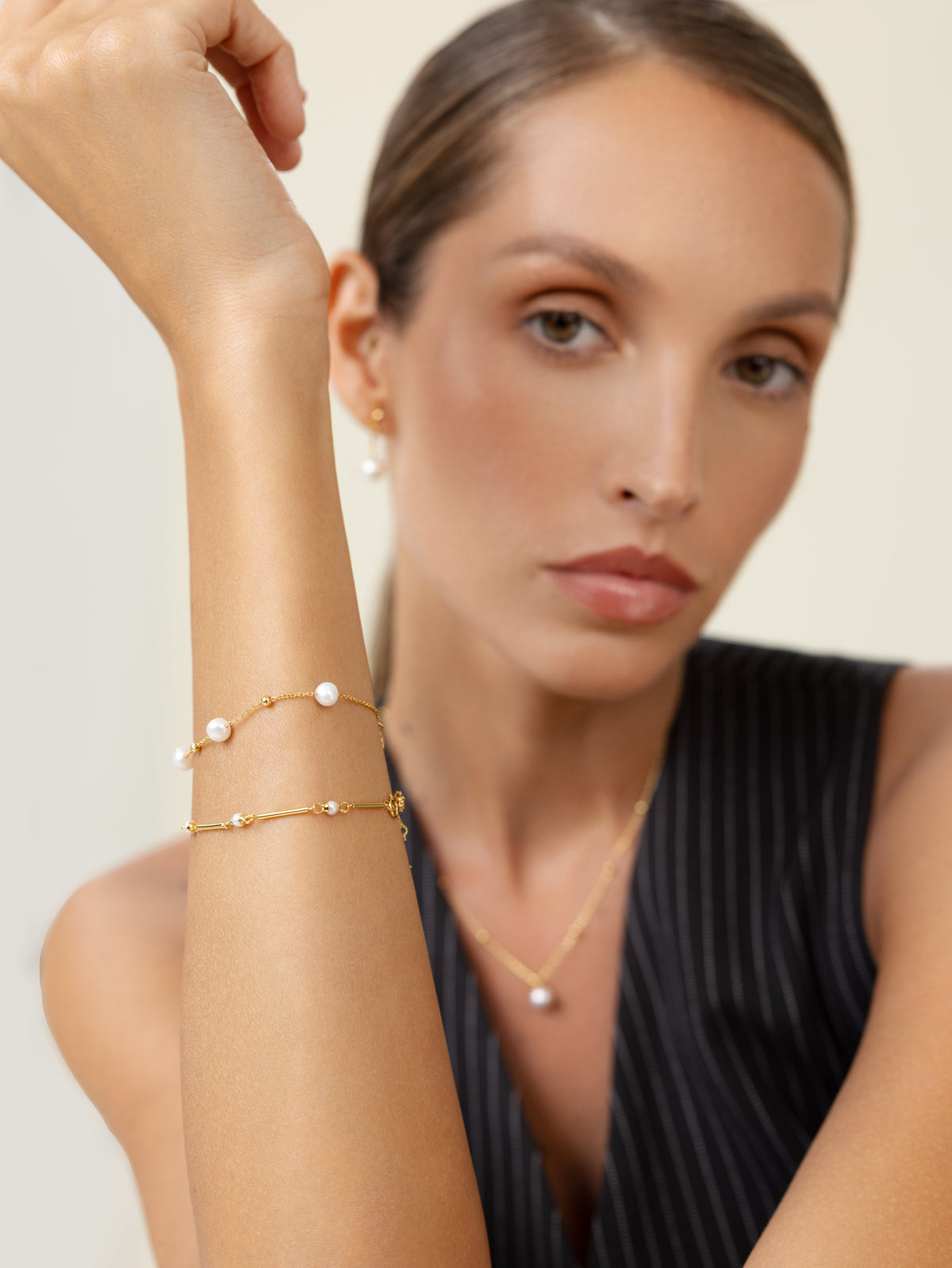 Woman in a pinstripe vest wearing stacked freshwater pearl station bracelets, a pearl pendant, and pearl earrings in 18k gold-plated sterling silver