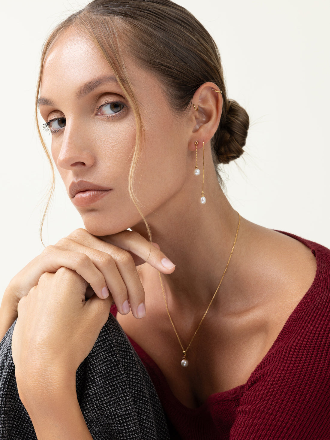 Model in a burgundy knit with a grey wool coat draped over her hand, wearing freshwater pearl drop earrings and a fine gold pearl pendant